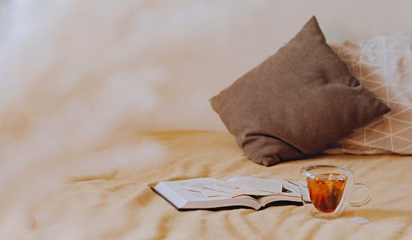 a cozy place to read at home with a book, cup of tea, pillows, and beige sheets